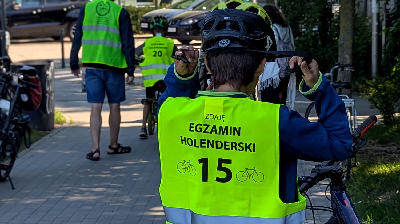 Stojący tyłem do kadru rowerzysta zakładający na głowę kask, ubrany w odblaskową kamizelkę. Na kamizelce napis Zdaję egzamin holenderski.